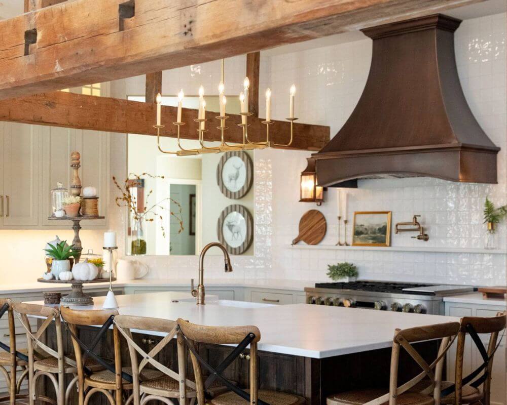 a rustic yet elegant kitchen with a blend of modern and traditional elements. The kitchen features a large island with a white countertop and a built-in sink. The island is surrounded by wooden chairs with woven seats and a crisscross back design. Above the island, there is a brass chandelier with exposed bulbs, adding a touch of vintage charm. The kitchen's focal point is a large, dark metal range hood above the stove, contrasting with the white tiled backsplash. The backsplash also includes a narrow shelf for displaying decorative items like cutting boards and framed art. To the right of the stove, there is a wall-mounted pot filler faucet, adding both functionality and style. The cabinetry is light gray with simple, classic hardware, blending seamlessly with the overall design.